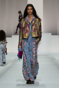 Model walks the runway in a gold metallic floral jacket over a blue striped floral jumpsuit, carrying a purple clutch.