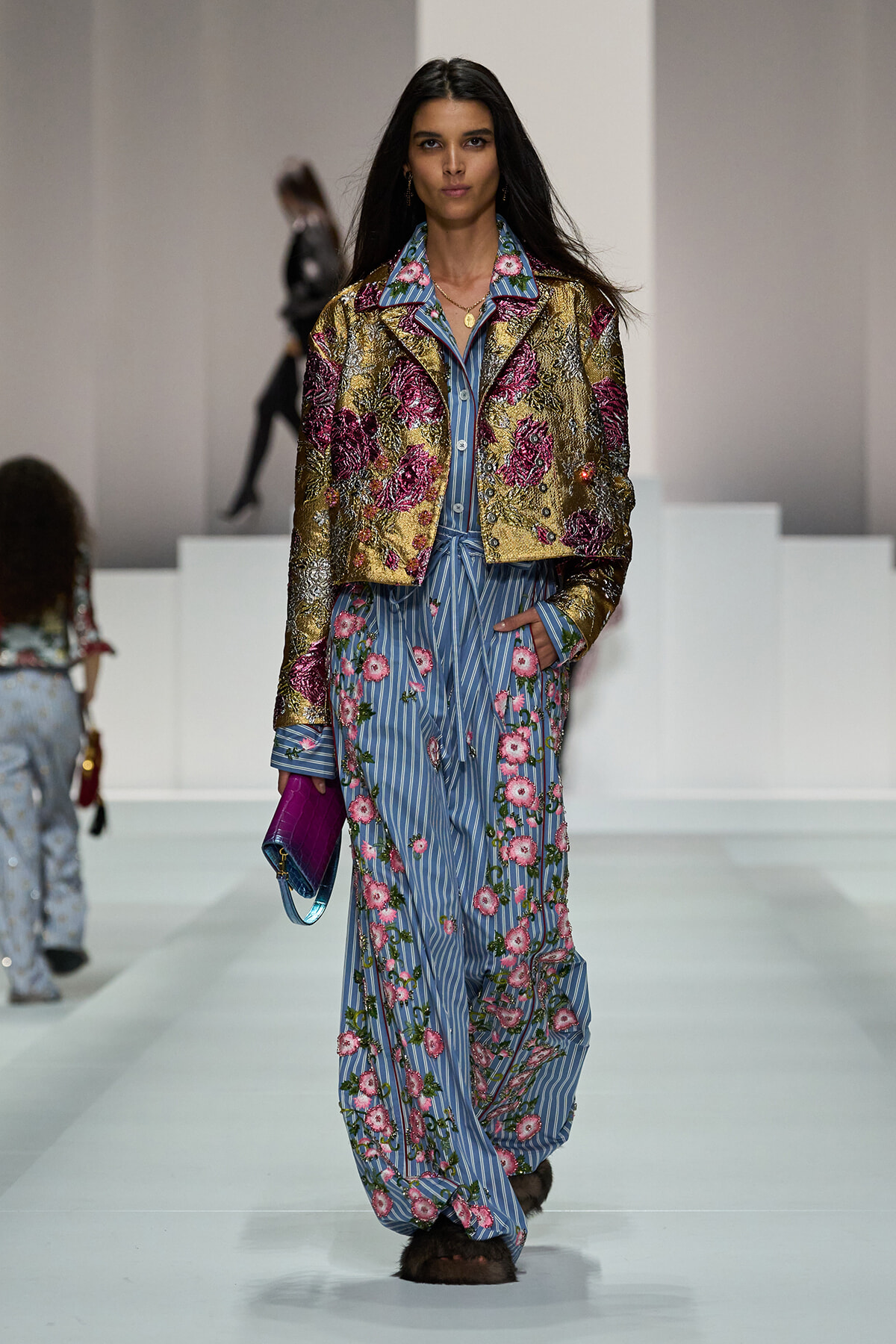 Model walks the runway in a gold metallic floral jacket over a blue striped floral jumpsuit, carrying a purple clutch.
