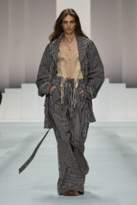 Model on a fashion runway wearing a gray pinstripe oversized coat and matching wide-leg pants over a beige corset, with a gold necklace.