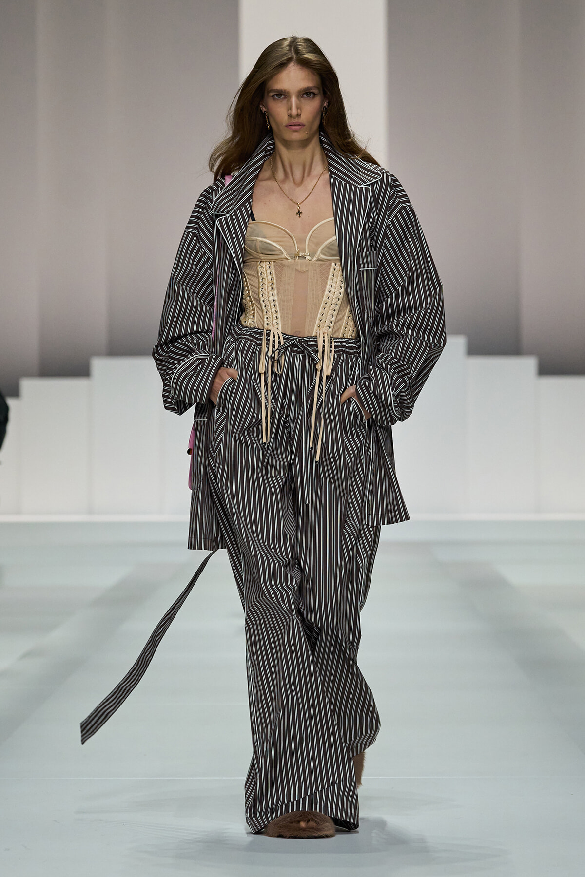Model on a fashion runway wearing a gray pinstripe oversized coat and matching wide-leg pants over a beige corset, with a gold necklace.