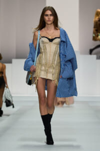 Model on a runway wearing a beige corset-style mini dress with lace trim and front lacing, paired with a blue striped oversized coat.