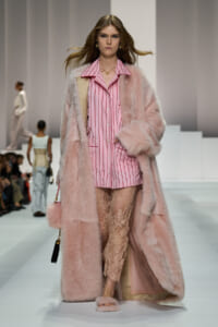 Model on a runway wearing a pink striped shirt, a long pink fur coat, lace pants, and pink fluffy slippers.
