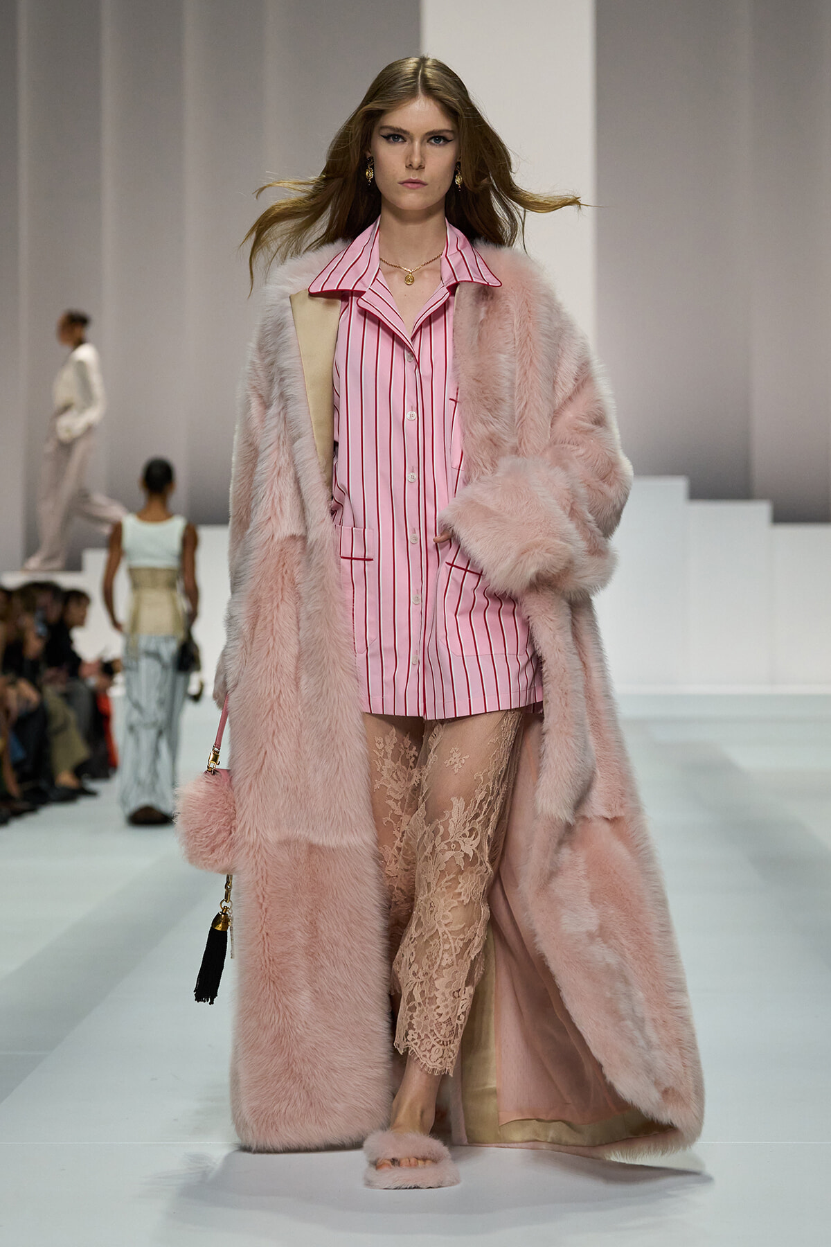 Model on a runway wearing a pink striped shirt, a long pink fur coat, lace pants, and pink fluffy slippers.