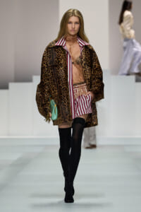 Runway model wearing a leopard-print coat over a pink striped shirt, black thigh-high socks, and a turquoise handbag.