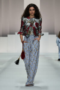 Model walking runway wearing a metallic floral jacket with red flowers and striped embroidered pants, carrying a red tassel bag.