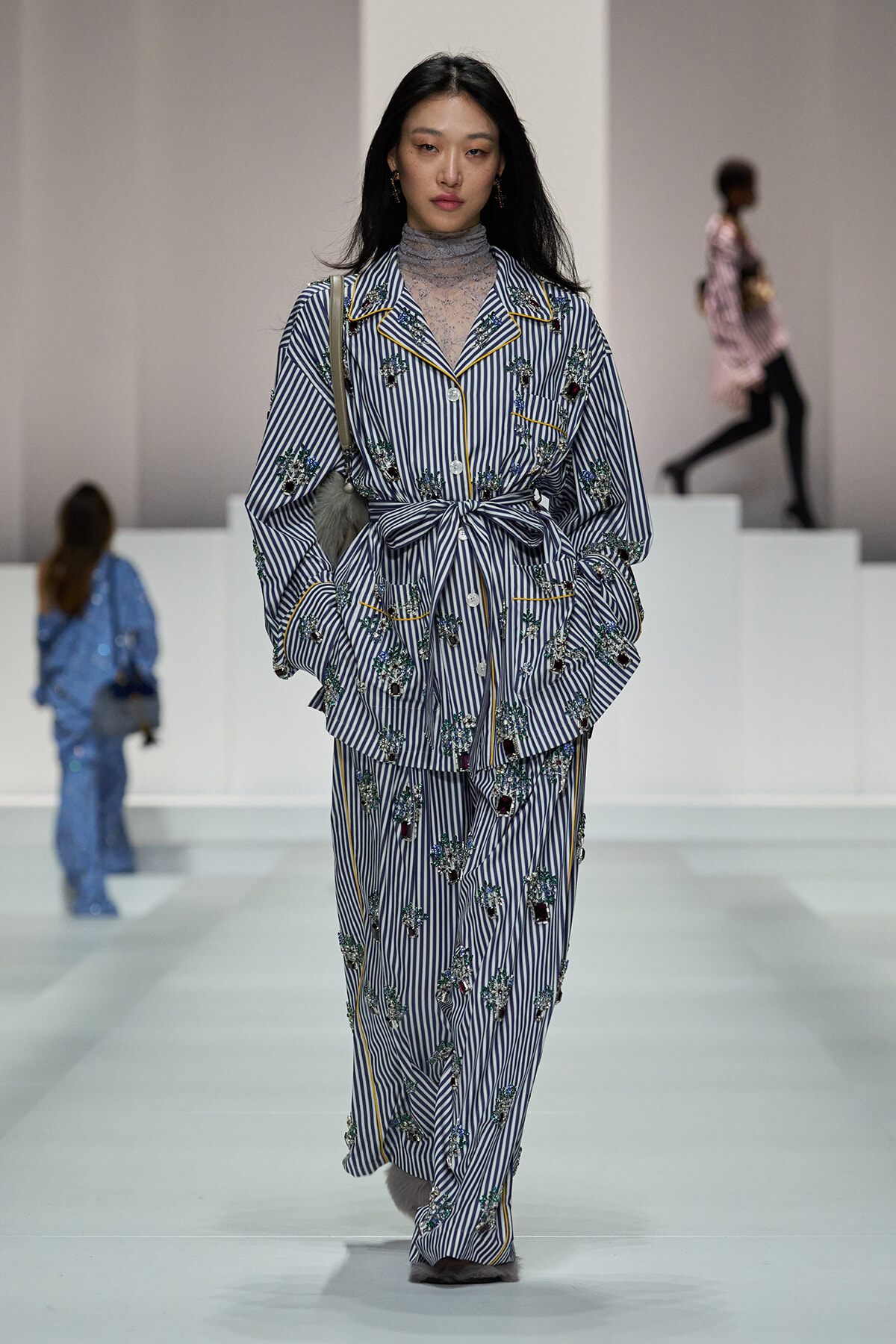 Model walking runway in striped floral-embellished suit with belt, lace turtleneck underneath.