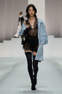 Runway model wearing a sheer black lace bodysuit under an oversized blue striped shirt, paired with black thigh-high socks and a black fringe bag.