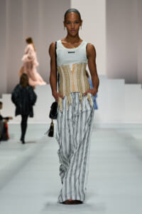 Model walking the runway in a beige lace-up corset over a pale blue sleeveless top, paired with wide white pants with vertical black stripes, carrying a small black handbag.