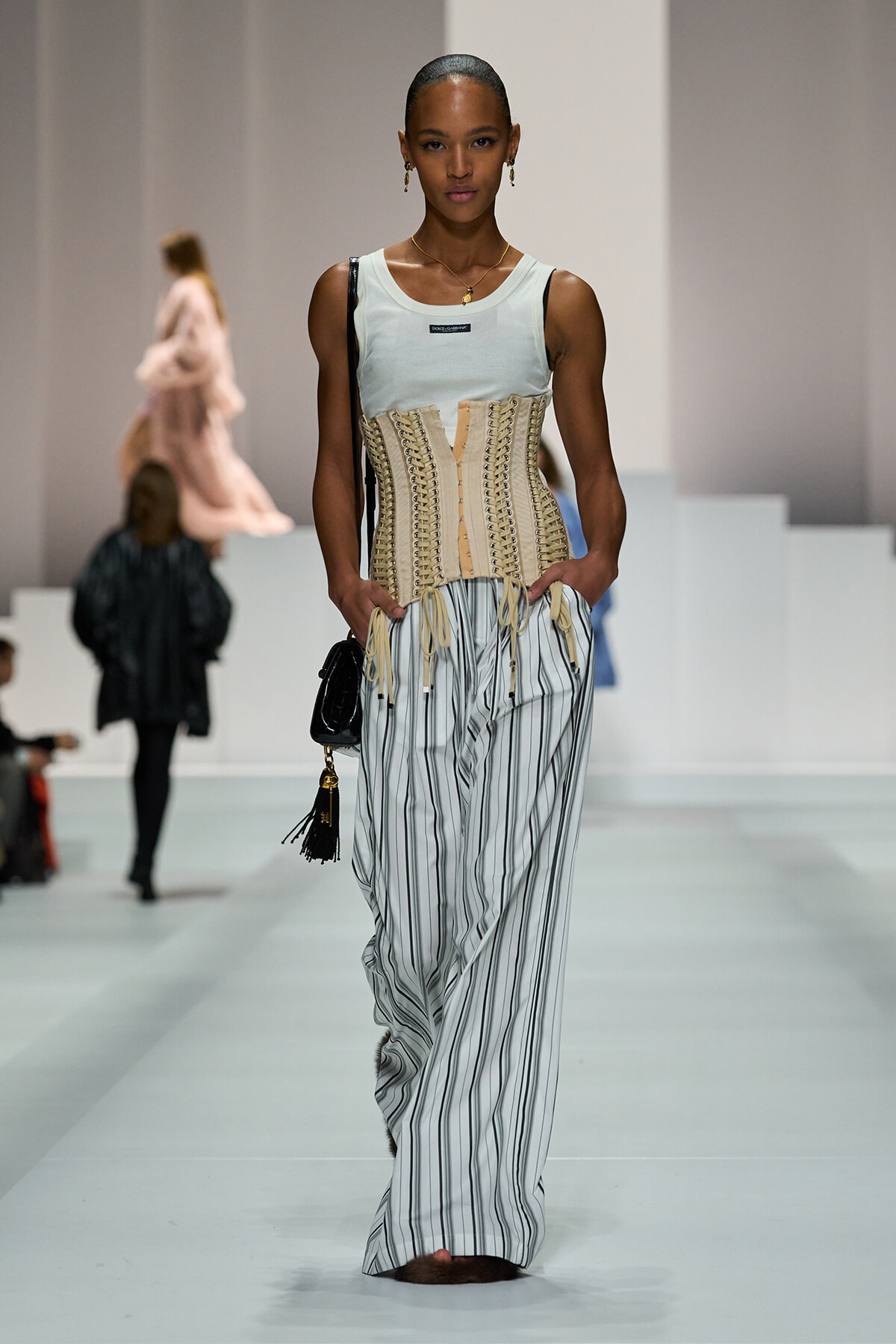 Model walking the runway in a beige lace-up corset over a pale blue sleeveless top, paired with wide white pants with vertical black stripes, carrying a small black handbag.