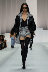 Model walking a fashion runway in a black leather jacket over a striped collared coat, black lace top, and striped shorts with thigh-high socks and heeled sandals.