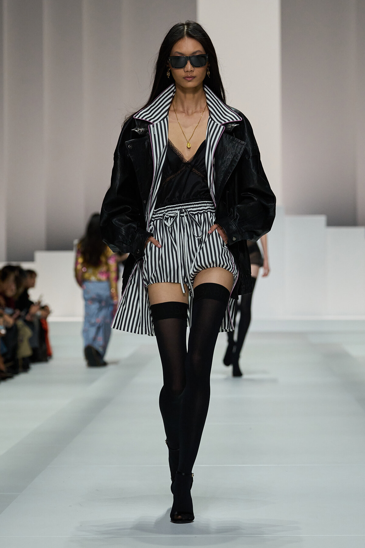 Model walking a fashion runway in a black leather jacket over a striped collared coat, black lace top, and striped shorts with thigh-high socks and heeled sandals.