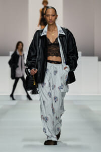 Model on a runway wearing a black leather jacket over a light-blue striped floral set with a black lace bralette and a black tassel bag showcased.