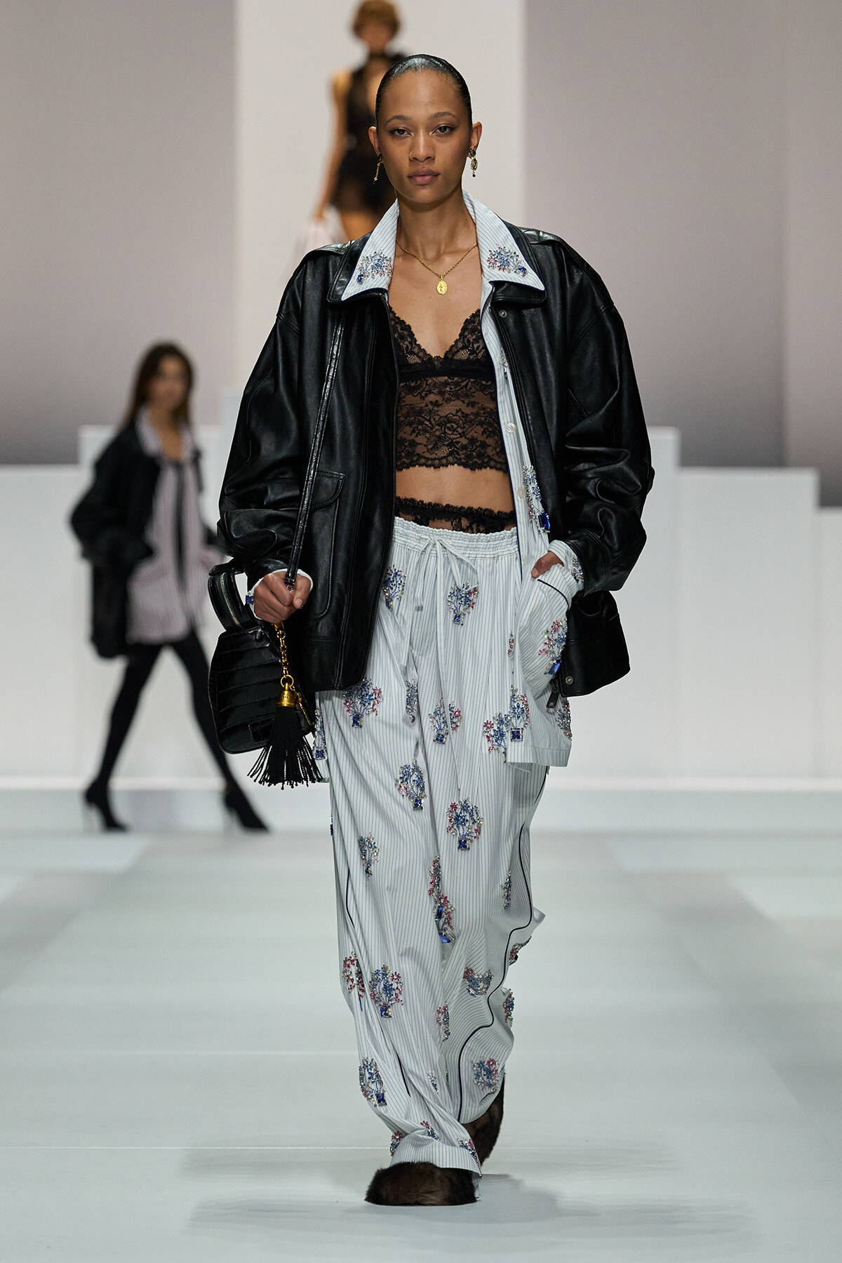 Model on a runway wearing a black leather jacket over a light-blue striped floral set with a black lace bralette and a black tassel bag showcased.