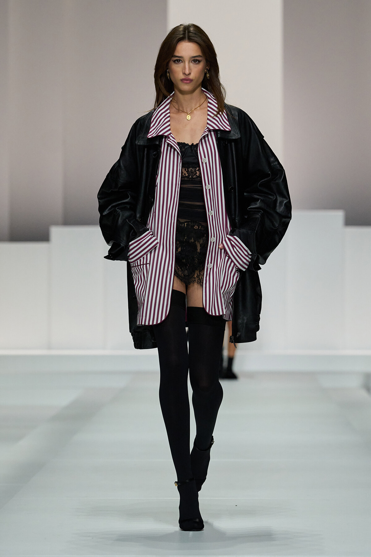 Model walking runway in oversized black leather coat over a maroon-and-white striped shirt and black lace dress, black tights and heeled boots.