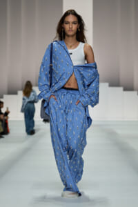 Model walking a fashion runway in a blue striped matching set with small floral motifs, a white sleeveless top, and the jacket casually draped off one shoulder; hands in pockets and fluffy white footwear add a relaxed vibe.