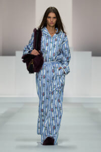 Model walking the runway in a blue-and-white striped floral loungewear set, carrying a burgundy fur bag.