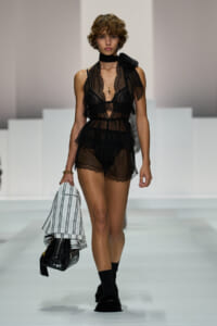 Model walks the runway in a sheer black lace romper with ruffle trim, paired with a black neck scarf and layered jewelry. She carries a striped blazer and a small black handbag, and wears black socks with fluffy black footwear at a fashion show.