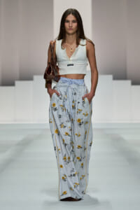 Model on a runway wearing a white sleeveless crop top and high-waisted striped floral pants, carrying a brown handbag.