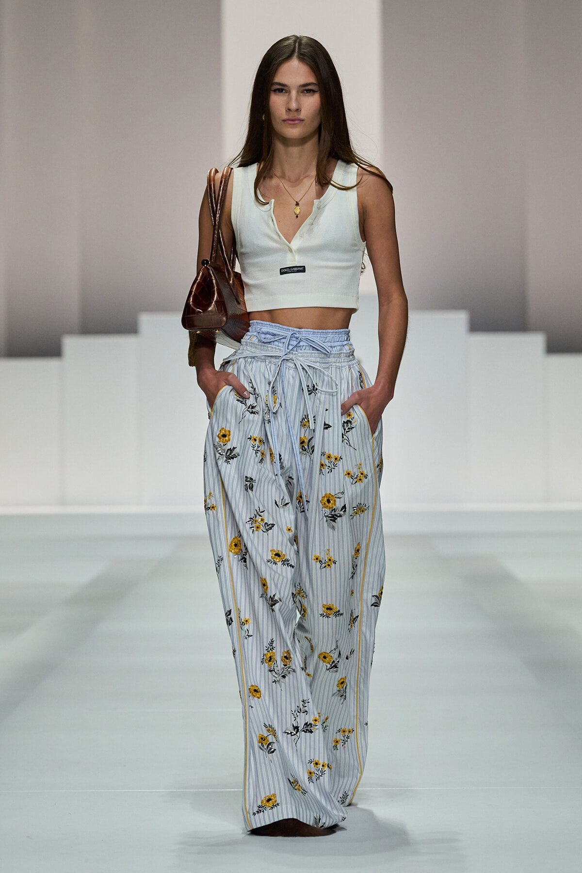 Model on a runway wearing a white sleeveless crop top and high-waisted striped floral pants, carrying a brown handbag.
