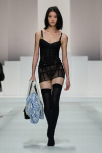 Female model walking a runway in a black lace bodysuit with sheer panels, thigh-high stockings, and a blue striped bag with white faux-fur trim in hand.