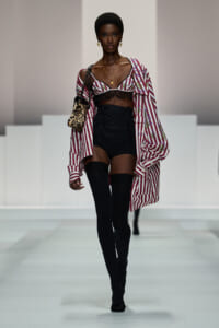 Model walking a fashion runway in a pink-and-white striped oversized shirt worn off one shoulder over a black lace bralette, black high-waisted shorts, and thigh-high socks, carrying a leopard-print bag with gold jewelry.