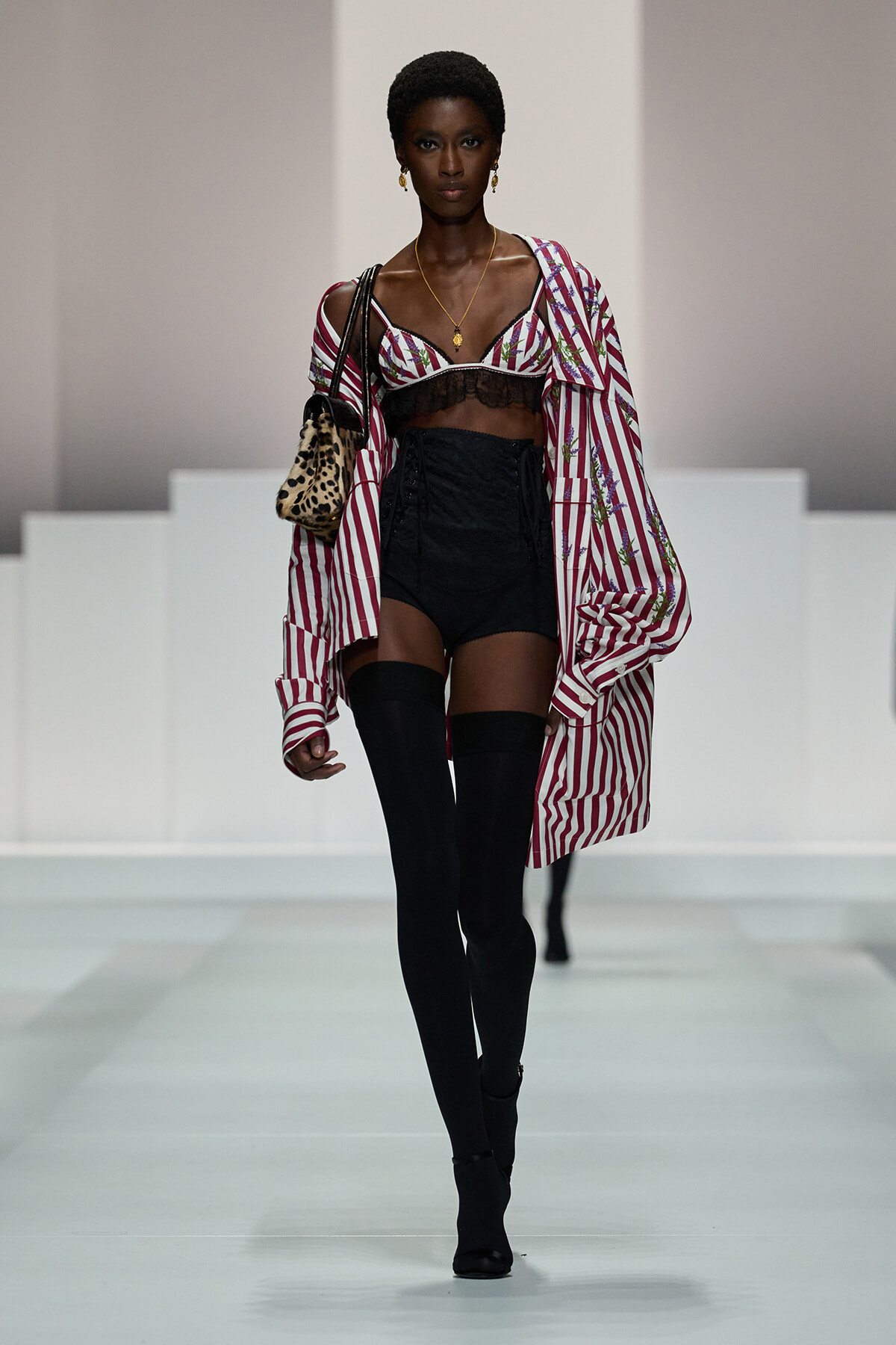 Model walking a fashion runway in a pink-and-white striped oversized shirt worn off one shoulder over a black lace bralette, black high-waisted shorts, and thigh-high socks, carrying a leopard-print bag with gold jewelry.