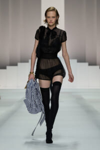 Model walking the runway in a sheer black blouse and high-waisted shorts with thigh-high stockings, carrying a blue-and-white striped bag.
