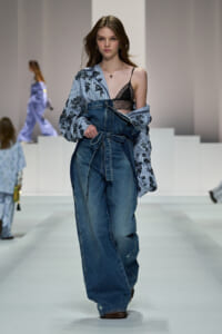 Model walking the runway in oversized denim overalls with a striped floral shirt draped over one shoulder, exposing a black lace bralette.