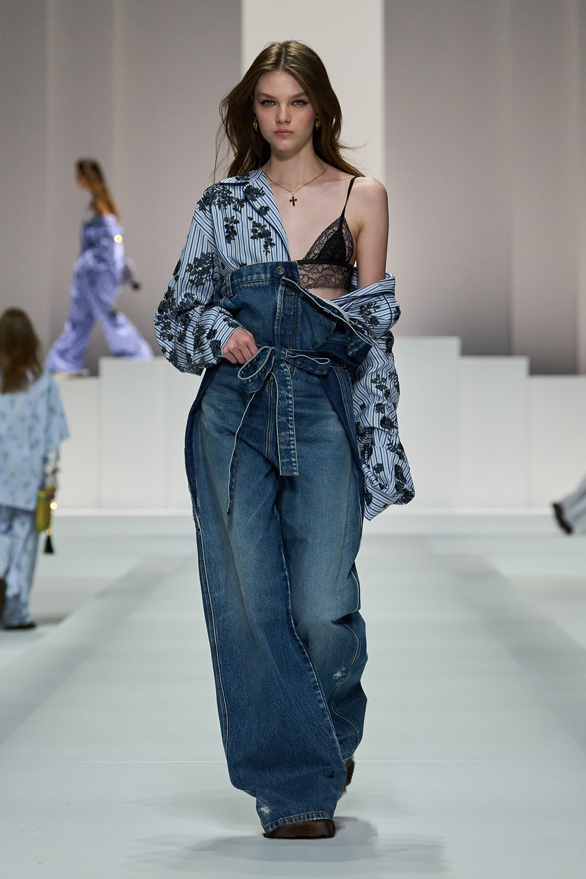 Model walking the runway in oversized denim overalls with a striped floral shirt draped over one shoulder, exposing a black lace bralette.