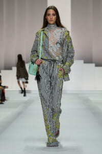 Model walking runway in black-and-white striped suit with yellow floral embroidery and a sheer lace turtleneck top; mint green handbag in left hand