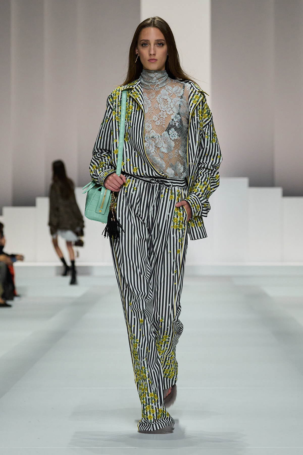 Model walking runway in black-and-white striped suit with yellow floral embroidery and a sheer lace turtleneck top; mint green handbag in left hand