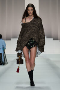 Model walks the runway in a loose brown knit poncho over a striped shirt, floral black shorts, and black socks, carrying a small brown handbag with a red tassel.