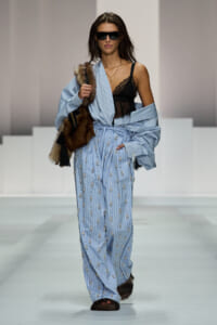 Runway model wearing light blue striped embellished jumpsuit with a fur stole, black lace top, and dark sunglasses.