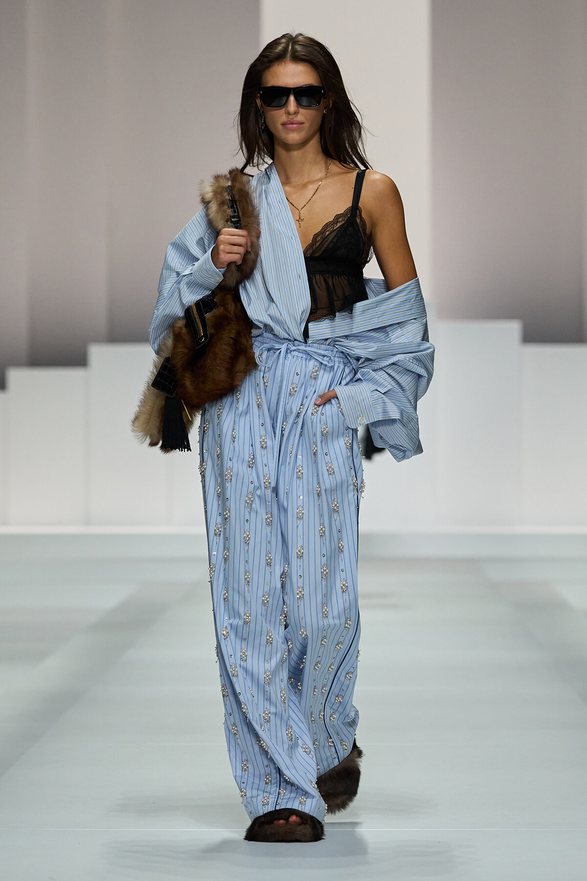 Runway model wearing light blue striped embellished jumpsuit with a fur stole, black lace top, and dark sunglasses.