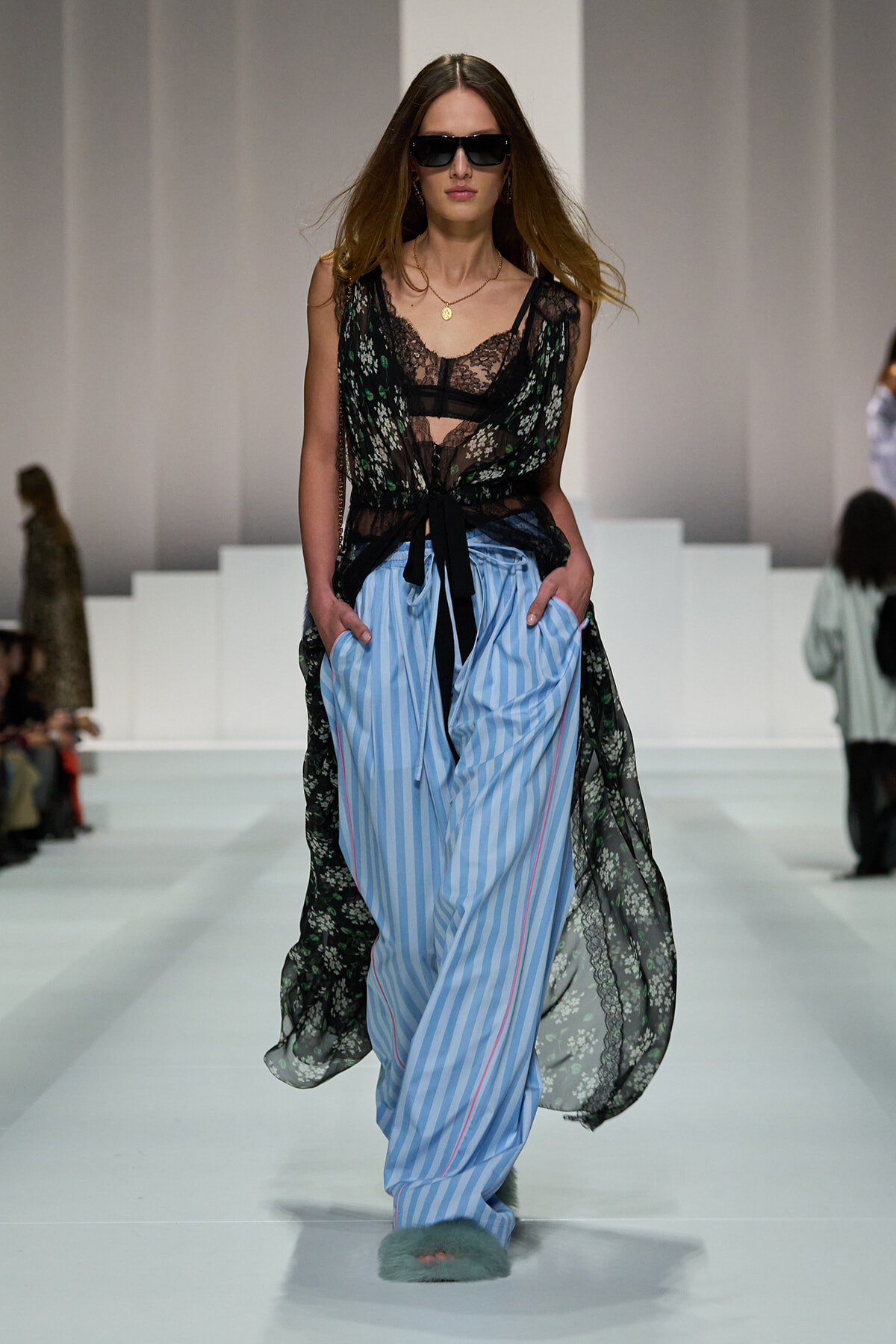 Model walking runway in black lace top over sheer floral overlay, tied at waist, paired with blue and white striped wide-leg pants and furry slippers, wearing dark sunglasses.