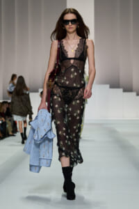 Model walking runway in sheer black lace dress with floral print, oversized sunglasses, and gold jewelry, carrying a light blue striped blazer.