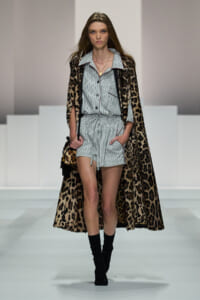 Model walking a fashion runway in a light striped matching two-piece romper under a full-length leopard-print cape, with black ankle boots and a small leopard bag.