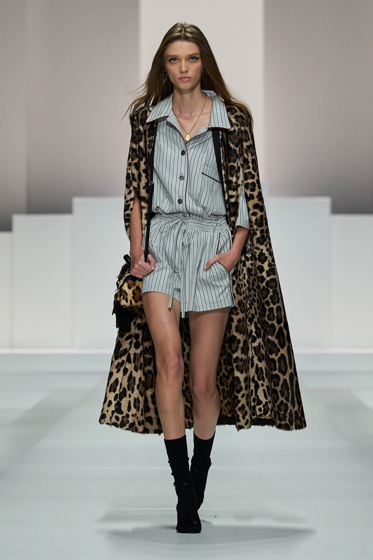 Model walking a fashion runway in a light striped matching two-piece romper under a full-length leopard-print cape, with black ankle boots and a small leopard bag.