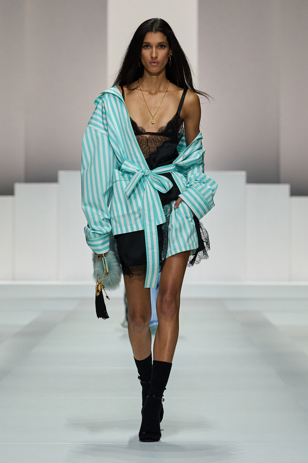 Model walks the runway in a turquoise and white striped oversized robe tied at the waist over black lace lingerie, accessorized with gold jewelry and a small handbag with tassels.