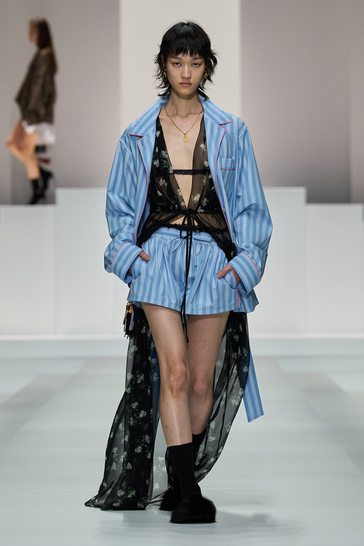 Model walks a runway in a light blue striped suit with matching shorts and an oversized blazer, worn over a sheer black lace top, with a long sheer floral cape; black socks and furry black footwear visible.