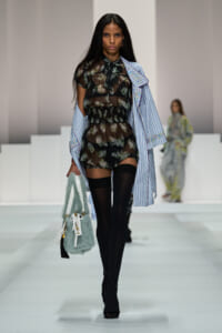 Model walking a runway in a sheer black floral romper with a light-blue striped shirt draped over her shoulders, black thigh-high socks, and a pale mint furry handbag.