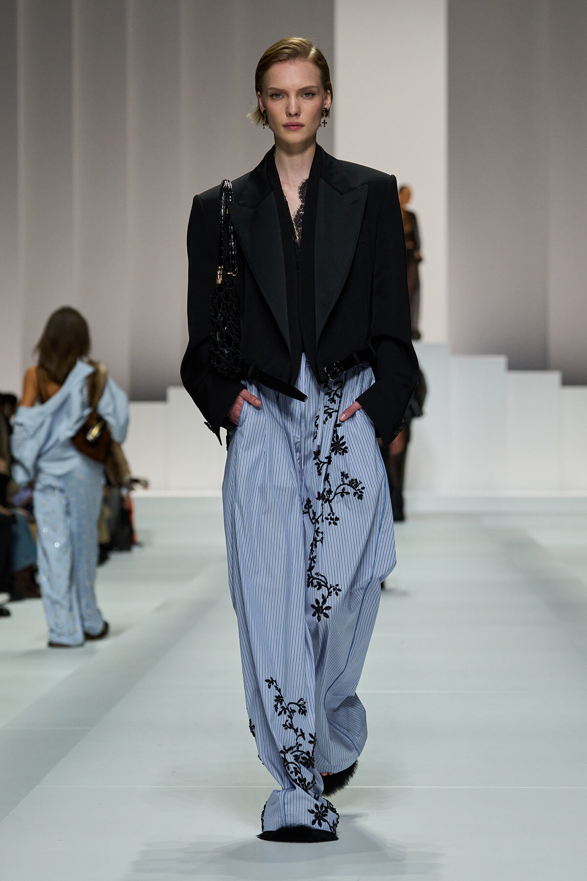Runway model in a black blazer over a lace top and blue striped pants with black floral embroidery, carrying a black bag.