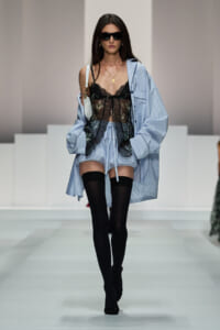 Model walks the runway in sheer black lace lingerie layered under a light blue striped oversized shirt, with black thigh-highs and sunglasses.