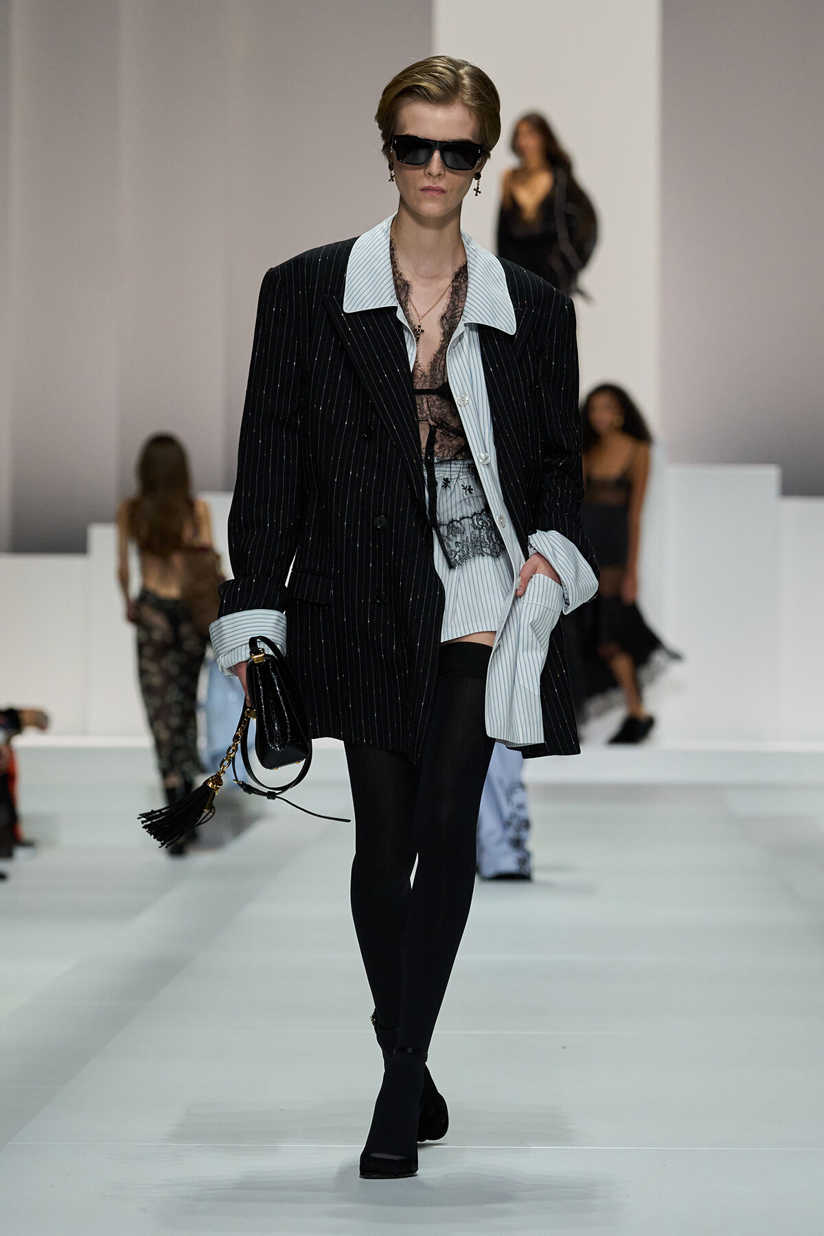 Model walks the runway in an oversized pinstripe blazer over a light blue striped shirt, black lace top, black tights, and black boots, accessorized with sunglasses and a tassel bag.