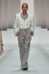 Model walking the runway in a white double-breasted blazer with striped lapels paired with high-waisted vertical-striped trousers, hands in pockets.