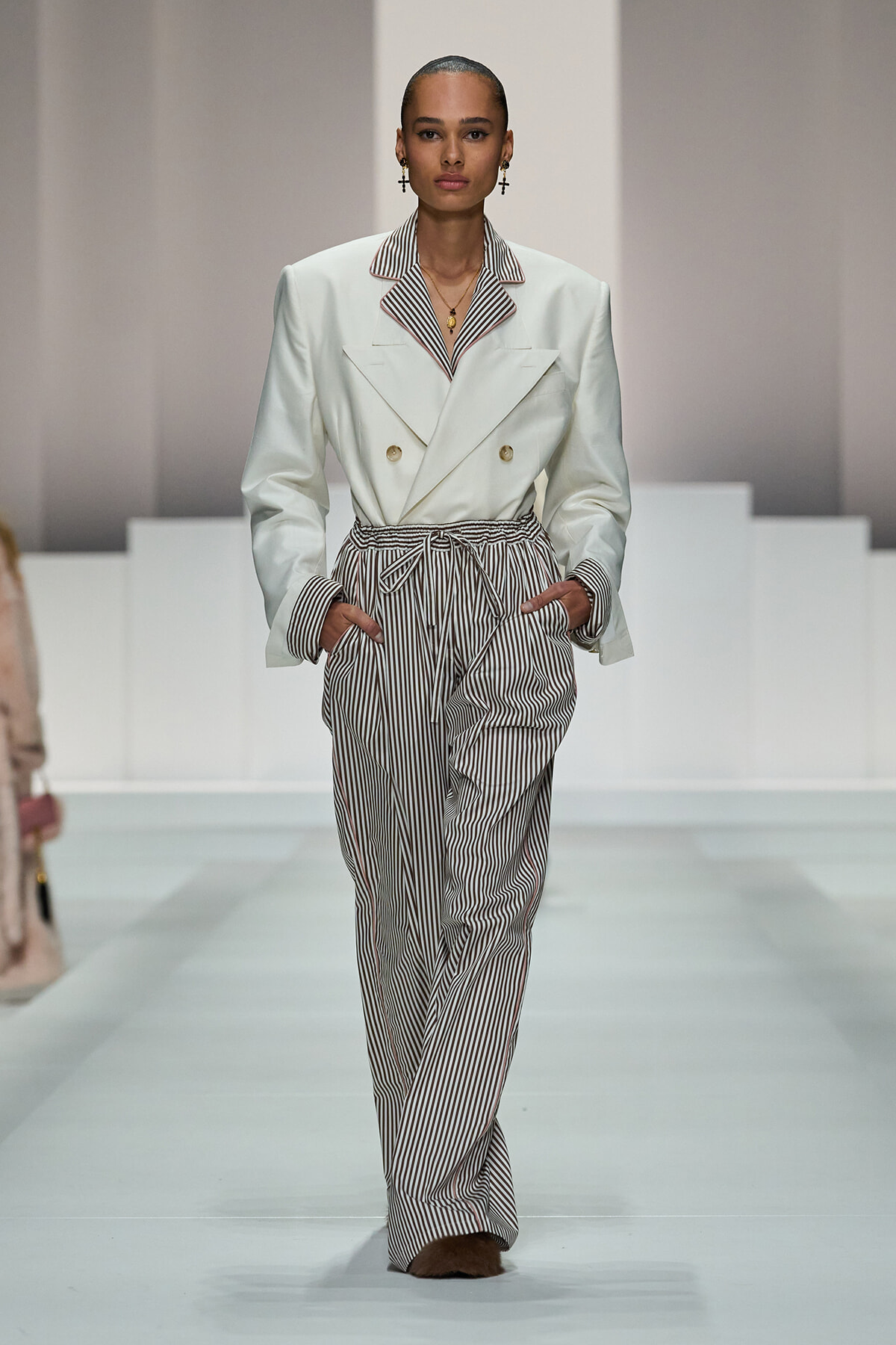 Model walking the runway in a white double-breasted blazer with striped lapels paired with high-waisted vertical-striped trousers, hands in pockets.