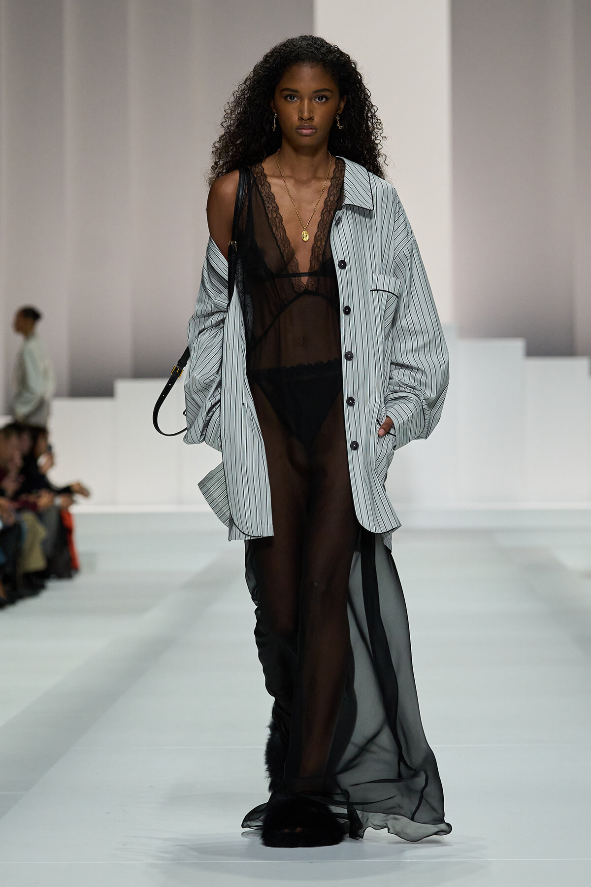 Model walks a fashion runway in a sheer black lace bodysuit under an oversized striped button-up shirt and a long sheer skirt, accessorized with layered gold necklaces and a small shoulder bag.
