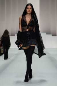 Runway model in sheer black lace top and black shorts with lacing, thigh-high stockings, and ankle boots, holding a small colorful clutch, cape draped over shoulders.