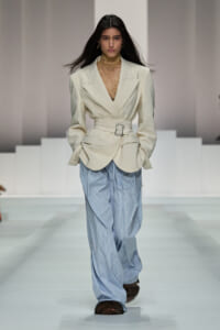 Model walking the runway in a cream tailored blazer with a wide belt over a sheer lace turtleneck and oversized light blue striped trousers, finished with brown fur-trimmed footwear.