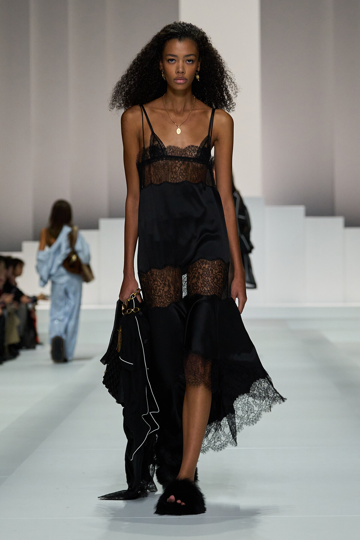 Model walking a runway in a black lace slip dress with sheer bodice and lace panels, carrying a black bag and wearing black fur slippers.